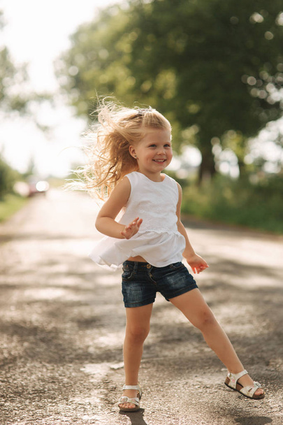 A cheerful adorable little girl with blond hair dances and jumps through the alley on a warm summer day - Foto, Imagem