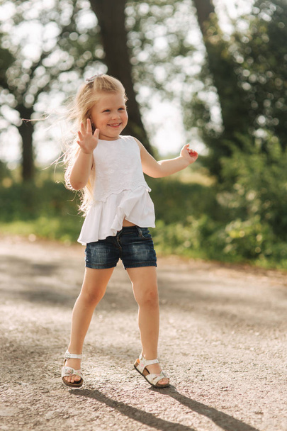 A cheerful adorable little girl with blond hair dances and jumps through the alley on a warm summer day - Valokuva, kuva