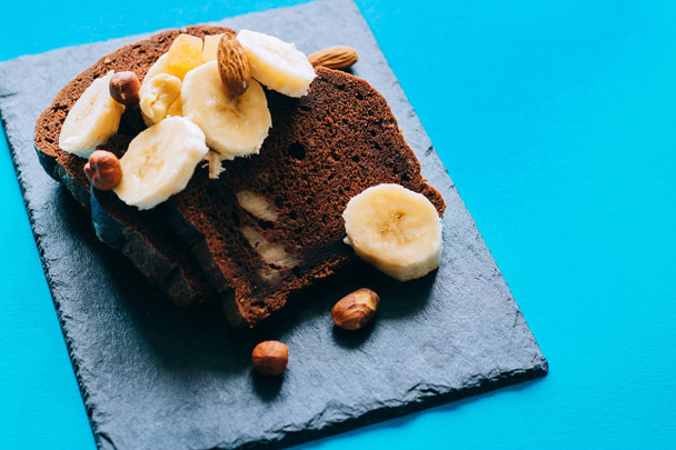 Chocolate banana bread on a black slate, with bananas and nuts for breakfast on blue background. - Foto, imagen