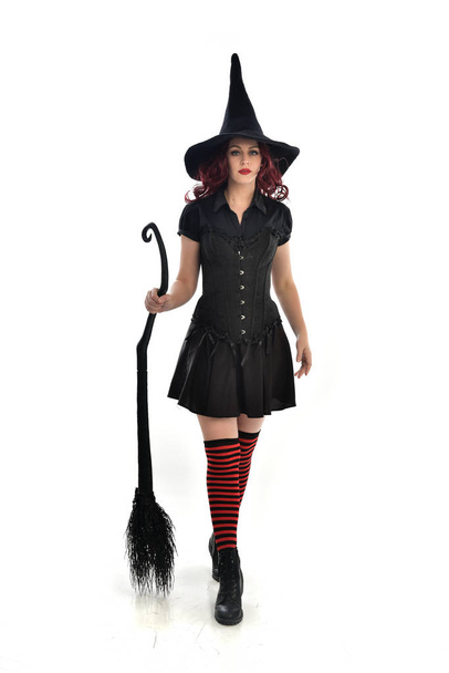 Ganzkörperporträt eines rothaarigen Mädchens mit schwarzem Hexenkostüm und spitzem Hut, das einen Besen in der Hand hält. Stehende Pose, isoliert auf weißem Studiohintergrund. - Foto, Bild
