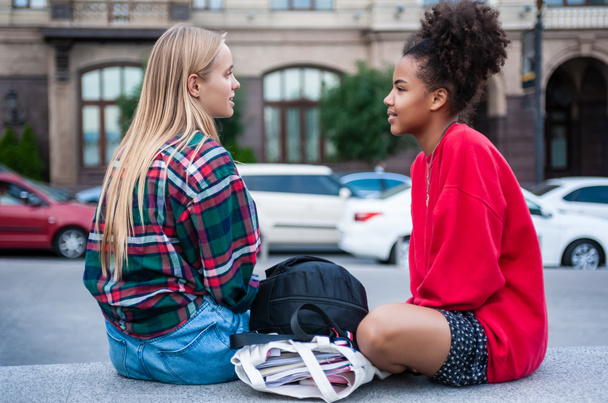 вид збоку на двох багатоетнічних дівчат, які сидять і дивляться один на одного на вулиці
 - Фото, зображення