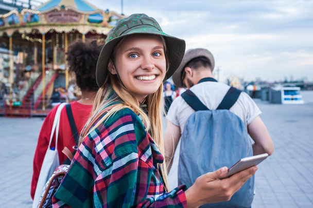 belle jeune femme tenant smartphone et souriant à la caméra tout en passant du temps avec des amis dans le parc d'attractions
 - Photo, image