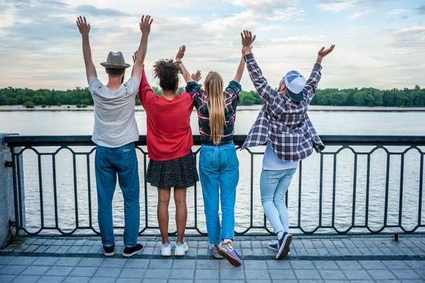 vista posteriore di quattro amici multietnici alzare le mani a riva del fiume
 - Foto, immagini