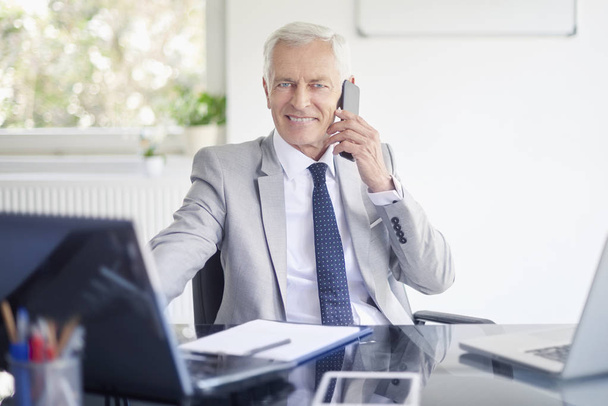 Εκτελεστικός ανώτερος επιχειρηματίας, μιλώντας στο smartphone ενώ εργασίας φορητό υπολογιστή στο γραφείο - Φωτογραφία, εικόνα