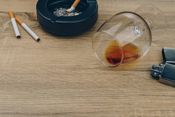 a glass of brandy, cigarettes, metallic lighter and black ceramic ashtray full of ashes with the cigarette butts on the wooden table - Photo, Image