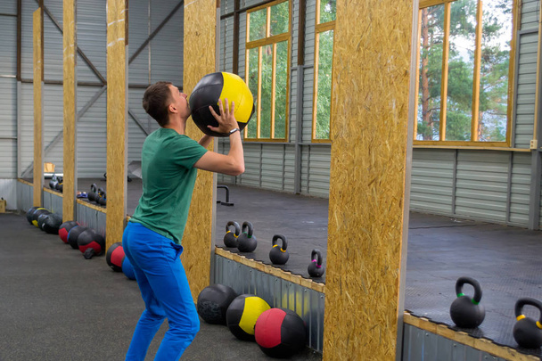ο τύπος σε ένα πράσινο μπλουζάκι και το μπλε παντελόνι τρένα στο γυμναστήριο, ρίχνει μια ιατρική μπάλα στον τοίχο, στο φόντο των γραμμών βάρη με πολύχρωμες λαβές και μια σύνθεση από αθλητικό εξοπλισμό - Φωτογραφία, εικόνα