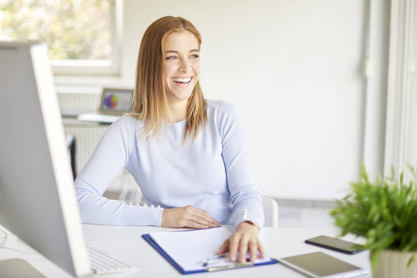 Smiling young businesswoman sitting in front of computer and posing in bright office - Foto, Bild