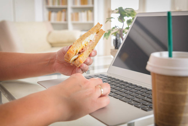 Woman eating a breakfast sandwich and drinking coffee while working with a laptop - Photo, image