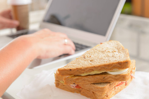 Woman eating a breakfast sandwich and drinking coffee while working with a laptop - Fotografie, Obrázek