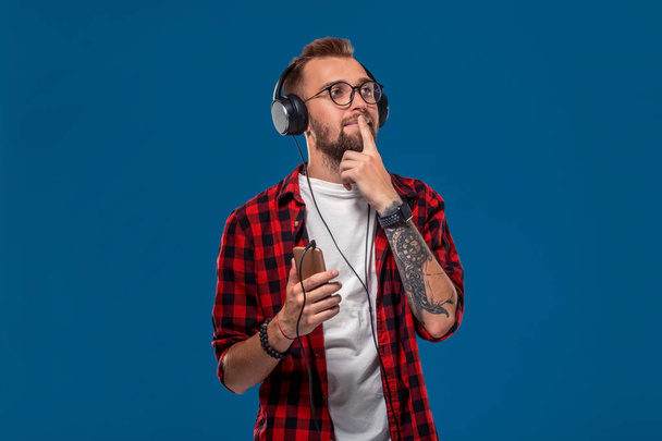 Handsome young stylish man in headphones holding MP3 Player and dancing while standing against blue background - 写真・画像