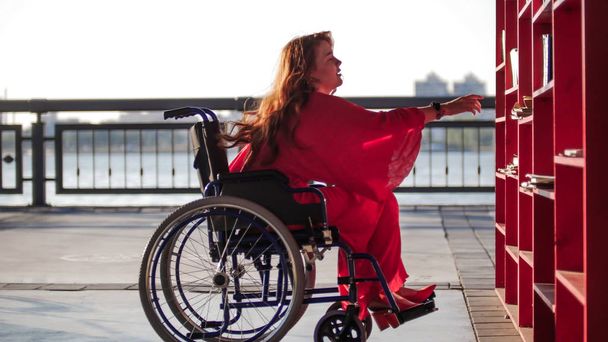schöne rothaarige Mädchen im Rollstuhl nimmt aus dem Regal Buch - Foto, Bild