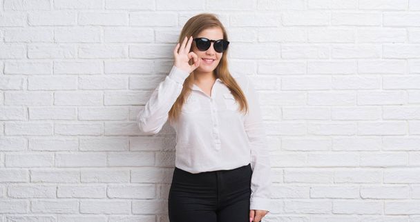 Young adult business woman standing over white brick wall wearing sunglasses doing ok sign with fingers, excellent symbol - Fotó, kép