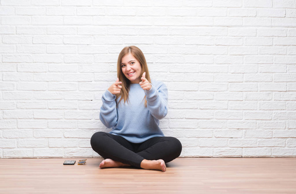 Young adult woman sitting on the floor in autumn over white brick wall pointing fingers to camera with happy and funny face. Good energy and vibes. - Photo, Image