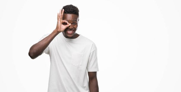 Молодой африканский американец в белой футболке делает жест с улыбкой на руке, глаза смотрят сквозь пальцы со счастливым лицом
. - Фото, изображение