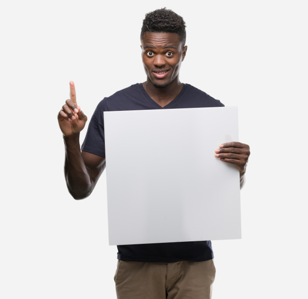 Young african american man holding a banner surprised with an idea or question pointing finger with happy face, number one - Foto, Imagen