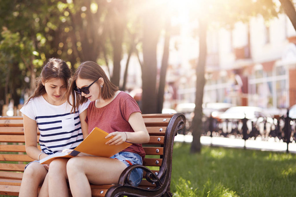 Two female students have attentive look in book, prepare for final exam at college, sit on bench in summer park, enjoy sunny weather, help each other with studying. People and communication. - Φωτογραφία, εικόνα