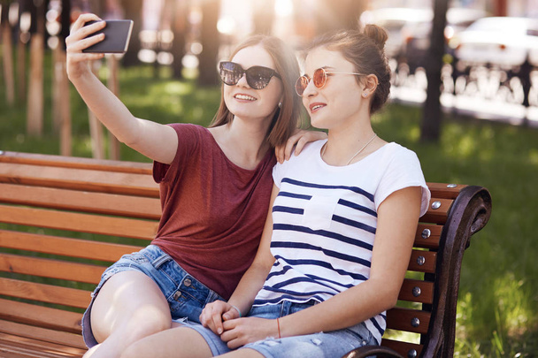 Positive female teenagers sit on wooden in bench in green park, pose for making selfie, look at smart phone, smile positively, do online translation, speak with friends, wears trendy sunglasses - Foto, Bild