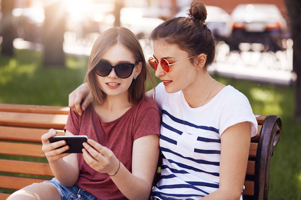 Best friends embrace while sit on bench in summer park, watch short videos on mobile phone, conncected to wireless internet, wear sunglasses, have good relationships. People and lifestyle concept - Foto, imagen