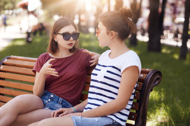Молодые женщины обсуждают свои планы на будущее после окончания школы, сидят вместе на скамейке запасных, приятно беседуют, наслаждаются летними каникулами и свободным временем. Женщины сплетничают о мальчиках. Концепция отдыха
 - Фото, изображение