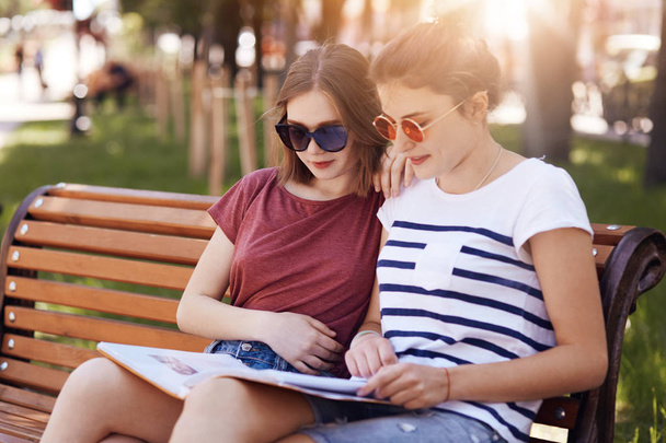 Outdoor shot of pleasant looking young female students being focused into scientific book, cramm for exam while st on bench in park and breath fresh spring air, prepare for session at university - Фото, изображение