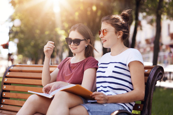 Outside shot of cheerful female friends, have gentle smiles, prepare for final exam together, sit on bench during hot summer day, being satisfied with something. People and lifestyle concept - 写真・画像