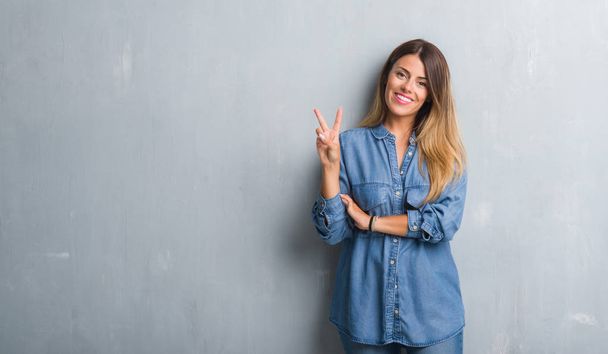 Молодая взрослая женщина за гранж-серой стеной в джинсовой одежде улыбается со счастливым лицом, подмигивая в камеру, делая знак победы. Номер два:
. - Фото, изображение