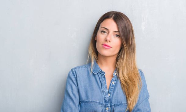 Молодая взрослая женщина на серой гранж-стене в джинсовой одежде с уверенным выражением на умном лице думает серьезно
 - Фото, изображение