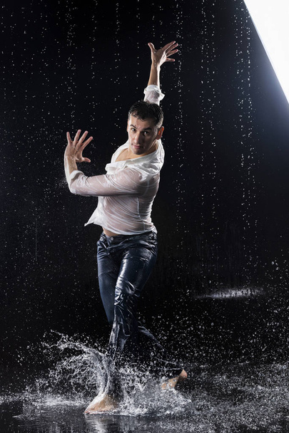 A young barefoot man wearing wet blue jeans and a white shirt expressively dances modern dances on the water under rainy waterdrops among the watersplashes. Isolated on a black background. Copy space. - Foto, imagen