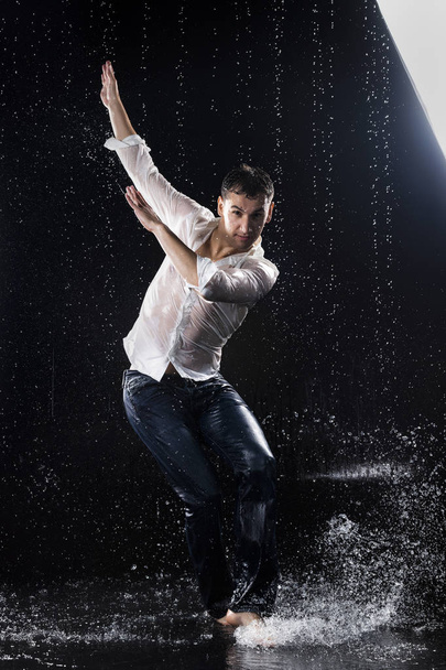 A young barefoot man wearing wet blue jeans and a white shirt expressively dances modern dances on the water under rainy waterdrops among the watersplashes. Isolated on a black background. Copy space. - Fotó, kép