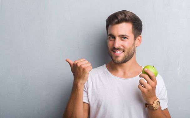 Όμορφος νεαρός άνδρας πάνω από την γκρίζα grunge τοίχο τρώει πράσινο μήλο κατάδειξης και δείχνει με αντίχειρα μέχρι την πλευρά με το χαρούμενο πρόσωπο χαμογελά - Φωτογραφία, εικόνα