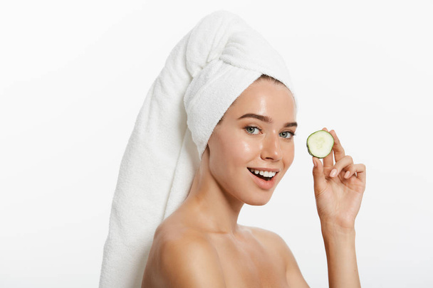Bliska portret uroda uśmiechający się piękny pół naga kobieta z ręcznik owinięty wokół jej włosy trzymając plasterki ogórka i patrząc na kamery na białym tle nad białym tle - Zdjęcie, obraz