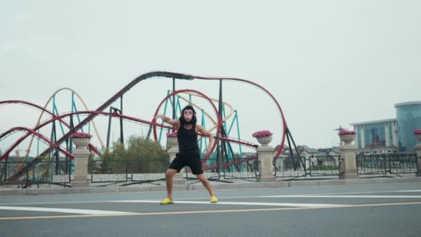 Hispanic man in monkey mask silly dancing in front of colorful roller coaster in amusement park with blue sky as background. Copyspace text - 映像、動画