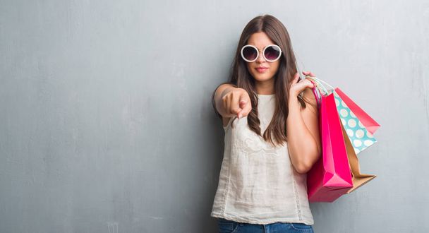 Młoda brunetka kobieta streszczenie szary mur gospodarstwa kolorowe torby na zakupy wskazując palcem do aparatu i do Ciebie, ręcznie zarejestrować, gest pozytywny i pewnie z przodu - Zdjęcie, obraz