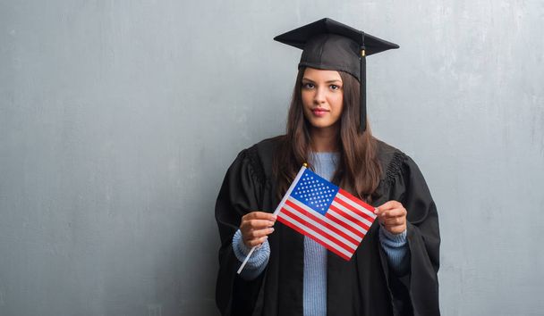スマートの顔は深刻な思考に自信を持って表現とアメリカの旗を保持している大学院の制服を着て不潔な灰色の壁を越えて若いブルネットの女性 - 写真・画像