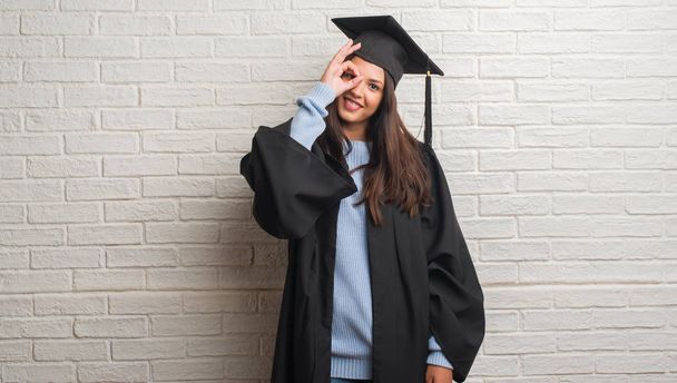 Mladá bruneta žena stojící nad bílou cihlová zeď postgraduální uniformě s šťastný obličej s úsměvem dělá ok znamení rukou na oko dívá skrze prsty - Fotografie, Obrázek