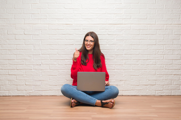 Ok işareti, başparmak ile parmak, mükemmel işareti yapıyor kocaman bir gülümseme ile laptop mutlu kullanarak beyaz tuğla duvarın üzerinden katta oturan genç esmer kadın - Fotoğraf, Görsel