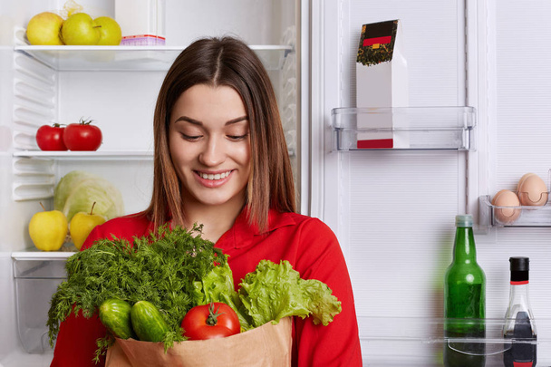 Portrait of happy woman with cheerful expression holds paper bag with tomatoes, cucmbers, dill and lettuce, comes from shop, going to make vegetable salad. People and healthy eatting concept - Photo, Image