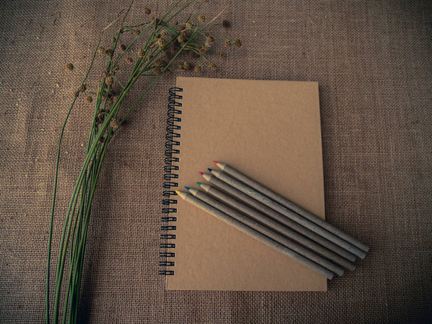 Vintage style. Organized desk with closed notebook, dry grass, pencils and burlap background - Photo, Image