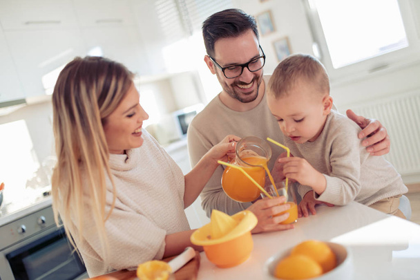 Εύθυμη νεαρή οικογένεια κάνει χυμό πορτοκάλι στην κουζίνα. - Φωτογραφία, εικόνα