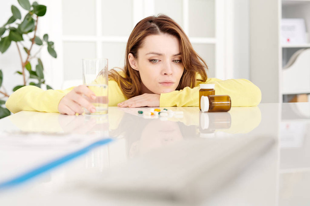 Portrait of sad young woman sitting at desk in doctors office and looking at assorted medication and pills - Foto, Imagen