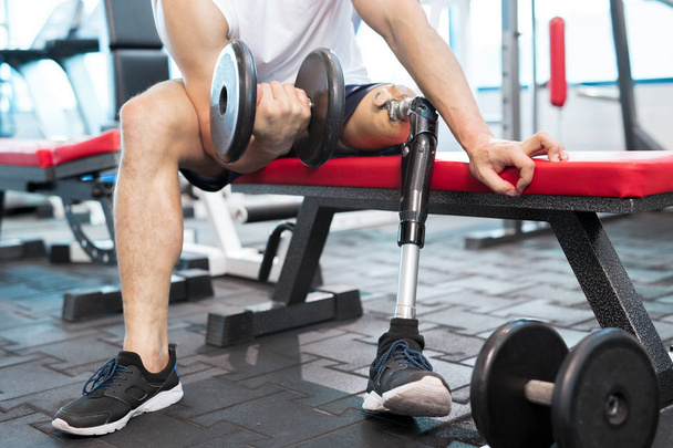 Retrato de sección baja de un hombre musculoso irreconocible con una pierna protésica haciendo ejercicio con una mancuerna sentada en el banco en el gimnasio moderno
 - Foto, Imagen
