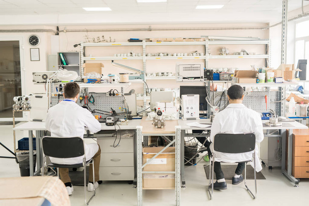 Taustakuva laboratoriotakkien kiireisistä insinööreistä, jotka istuvat pöydän tuoleilla ja kokoavat tai korjaavat mittauslaitetta manometrityöpajassa
 - Valokuva, kuva