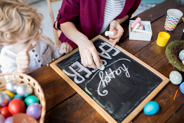 Zblízka mladé ženy psaní Veselé Velikonoce na tabuli zároveň ručně vyráběné ozdoby doma - Fotografie, Obrázek