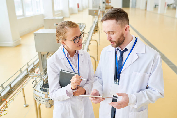 Handsome bearded technologist wearing white coat showing statistic data concerning production quantity to female colleague with help of digital tablet while standing at spacious dairy factory. - Foto, Imagen