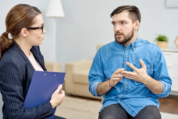 Depressed patient wearing denim shirt having face to face problem discussion with highly professional psychologist after completion of group therapy session, interior of cozy office on background - Photo, Image