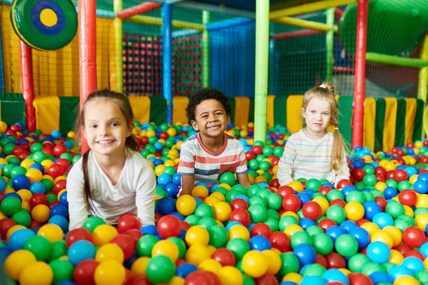 Красочный портрет трех счастливых маленьких детей в яме шаров, счастливо улыбающихся в камеру, веселясь в детском игровом центре
 - Фото, изображение