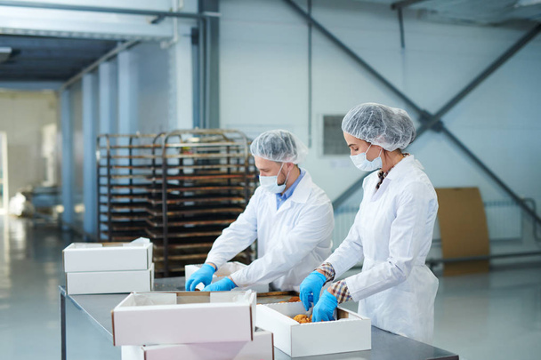 Travailleurs d'usine de confiserie en couches blanches mettant la pâtisserie prête dans des boîtes en papier
.  - Photo, image