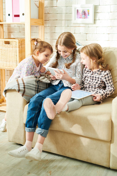 Три маленькие сестры весело проводят время, смотря свой любимый мультфильм на цифровом планшете, интерьер уютной гостиной освещается солнечными лучами на заднем плане
 - Фото, изображение