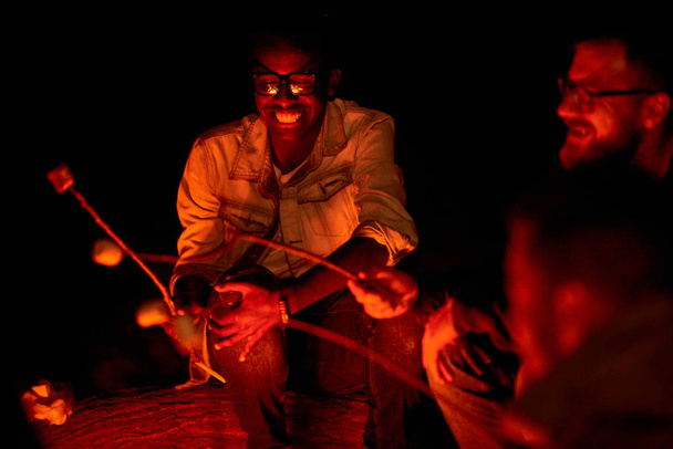 Wesoły zadowolony Afryki młodzieńca w okularach pieczenia Zefir podczas rozmowy do bliskich przyjaciół podczas nocy camping - Zdjęcie, obraz
