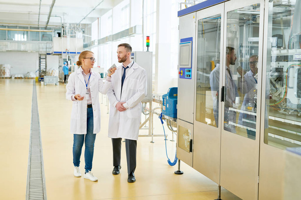 Pretty fair-haired entrepreneur wearing white coat holding smartphone in hand while giving tour around production department of dairy plant for bearded inspector - Photo, Image